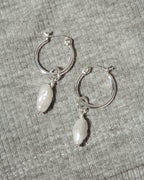 A pair of silver hoop earrings with dangling white Keshi pearls, displayed on a textured gray fabric background.