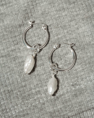 A pair of silver hoop earrings with dangling white Keshi pearls, displayed on a textured gray fabric background.