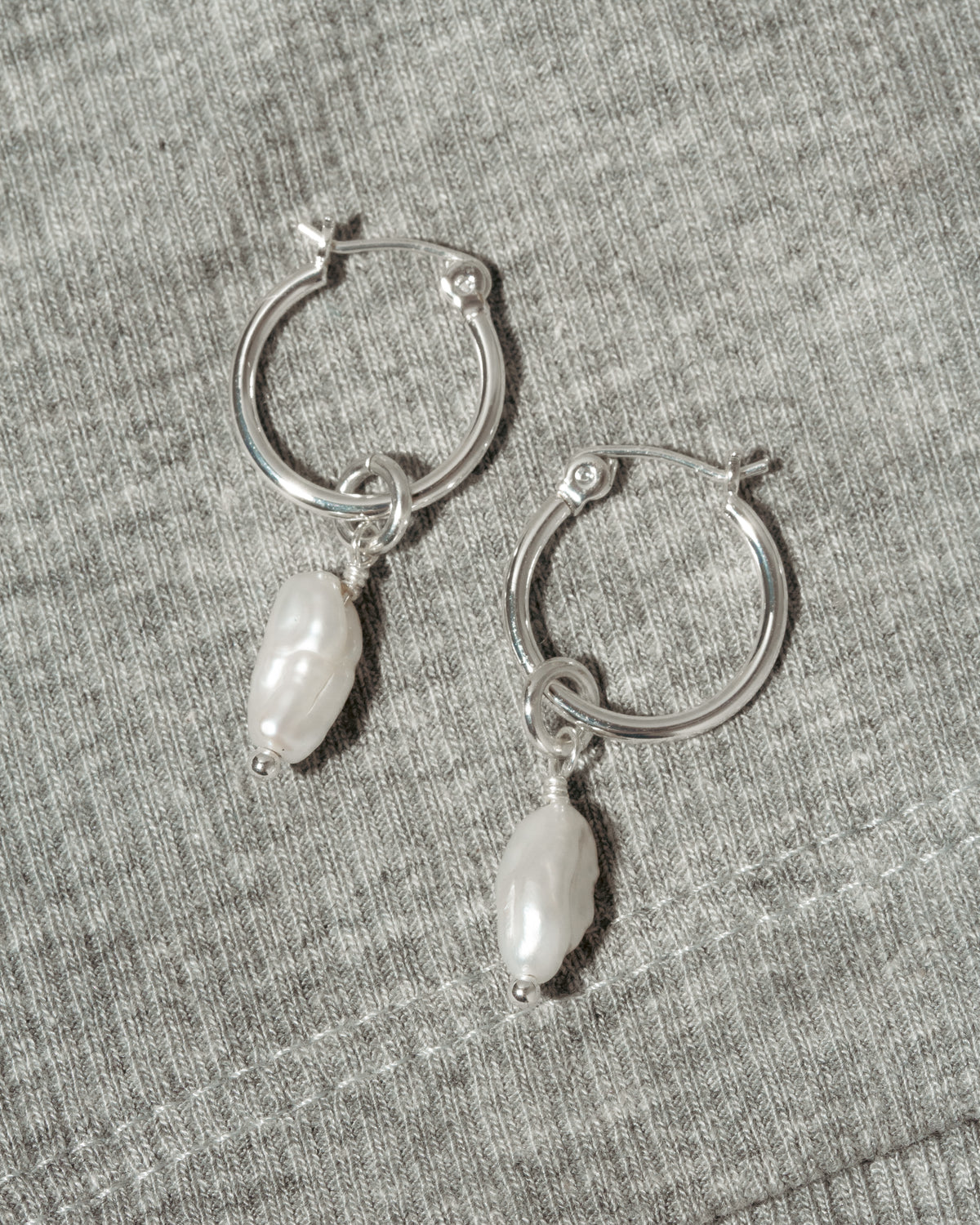 A pair of silver hoop earrings with dangling white Keshi pearls, displayed on a textured gray fabric background.