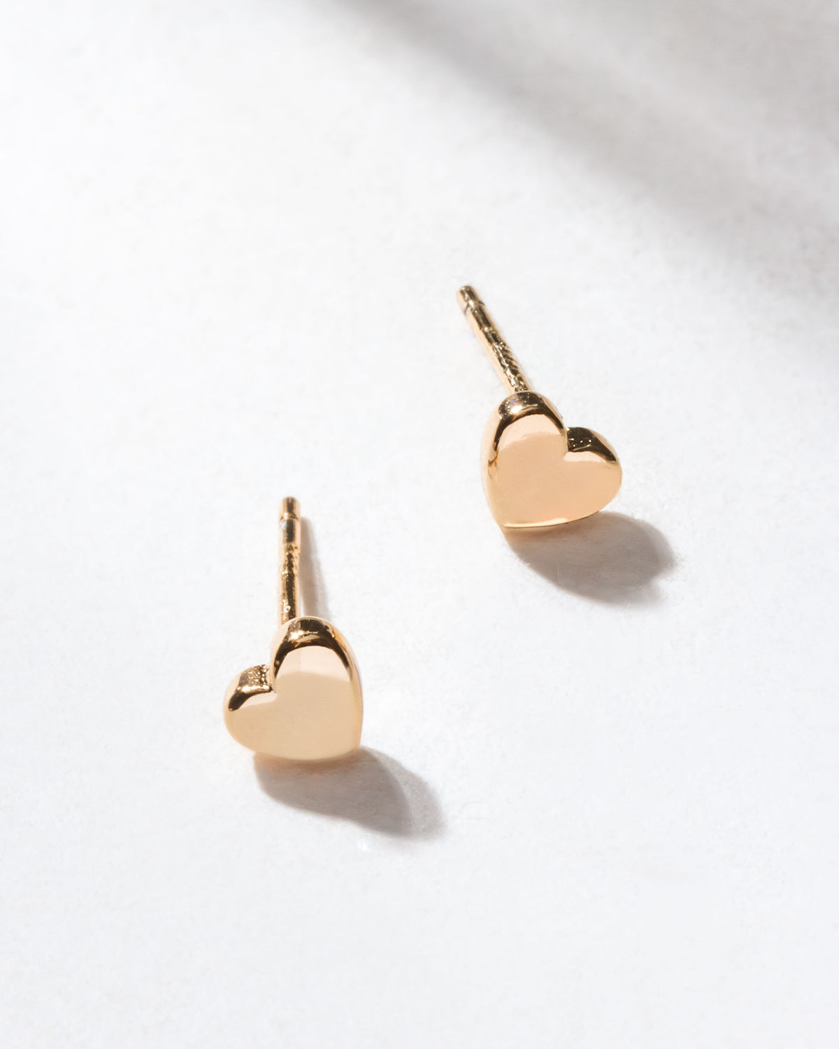 Two gold heart stud earrings are displayed on a white surface.