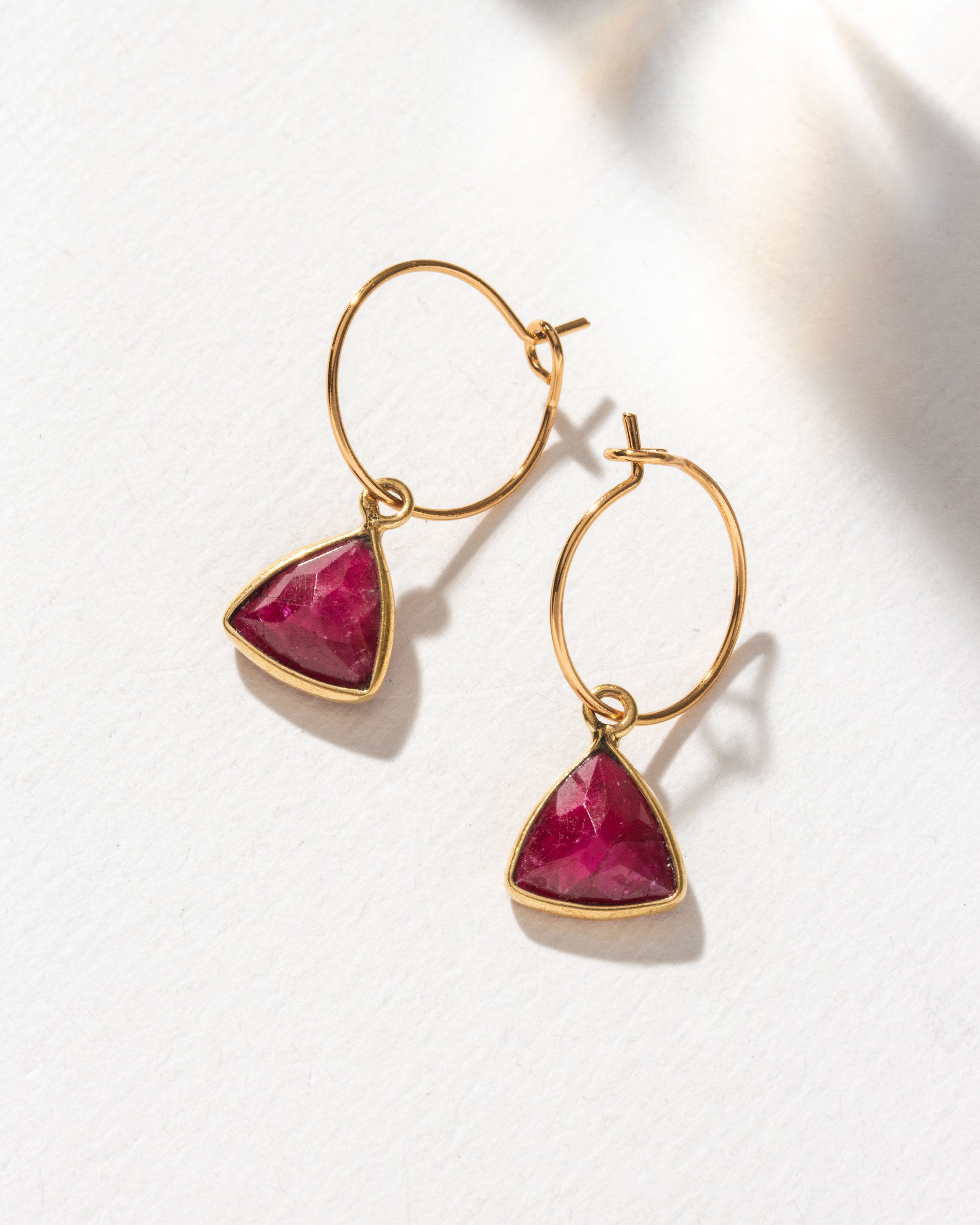 Gold mini hoops with a triangular Ruby gemstone charm are displayed.