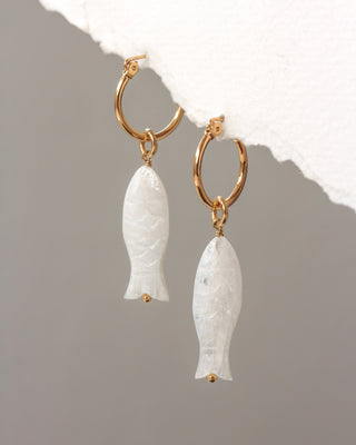 Gold hoop earrings with white stone fish pendants on a textured surface