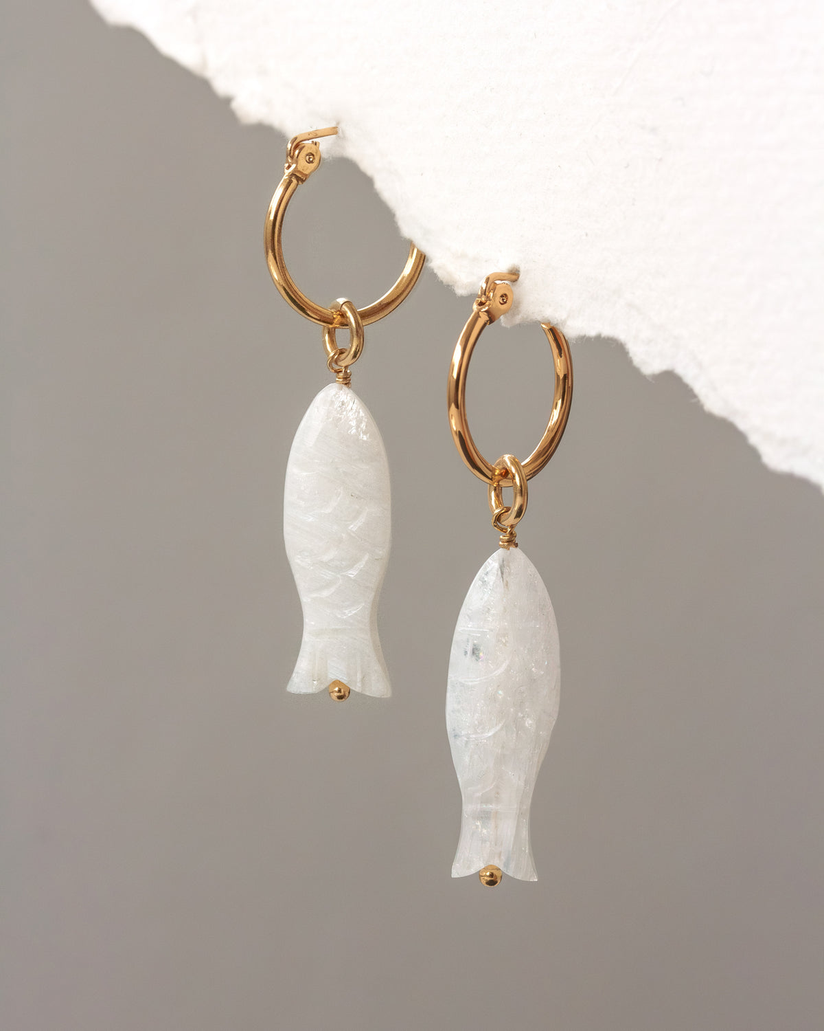 Gold hoop earrings with white stone fish pendants on a textured surface