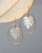 Gold hoop earrings with mother-of-pearl leaf-shaped cutouts hanging on a leaf