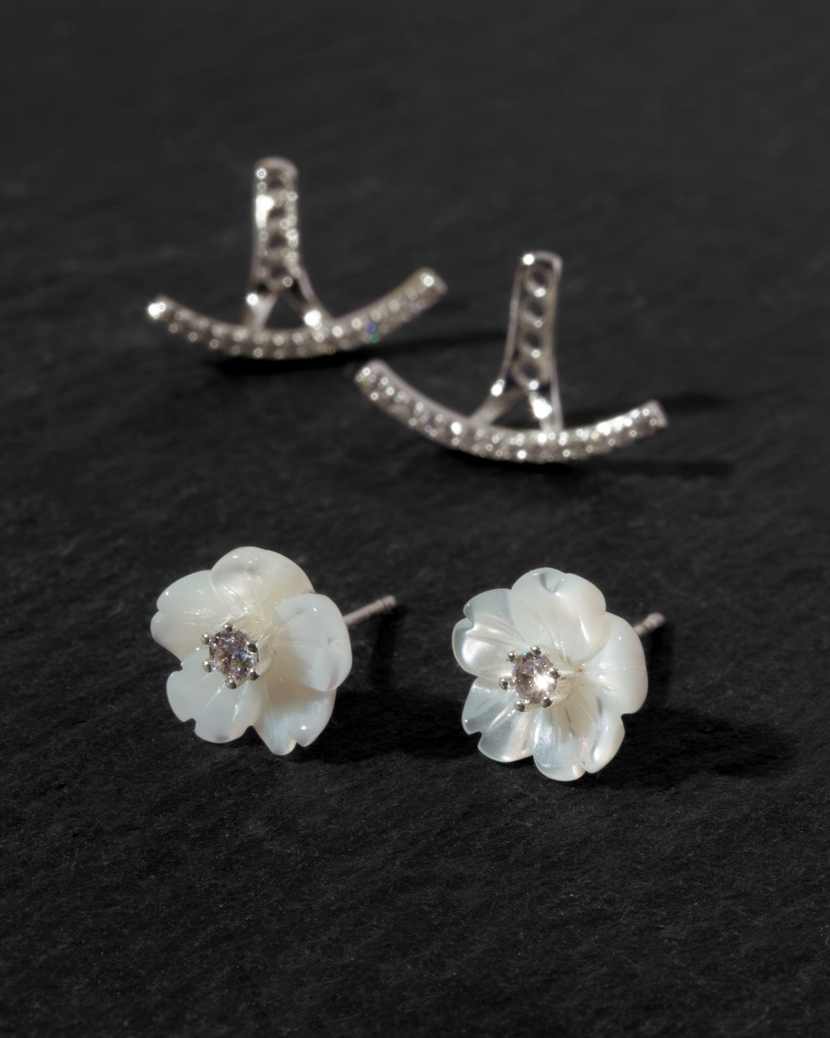 A pair of white mother-of-pearl flower stud earrings with clear CZ centers and a matching clear cz front back earring jacket, arranged on a dark textured background.