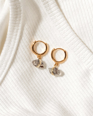 A pair of gold huggie hoop earrings with small, rough, clear Herkimer Quartz gemstone dangles, displayed on a textured white ribbed knit fabric.