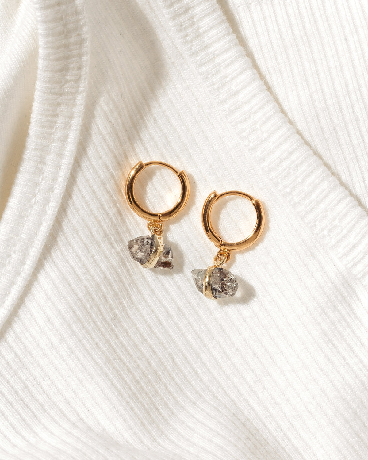 A pair of gold huggie hoop earrings with small, rough, clear Herkimer Quartz gemstone dangles, displayed on a textured white ribbed knit fabric.