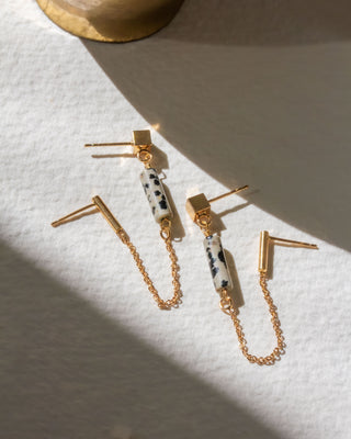 Gold double pierced earrings with cylindrical Dalmatian Jasper charms on a white textured surface.