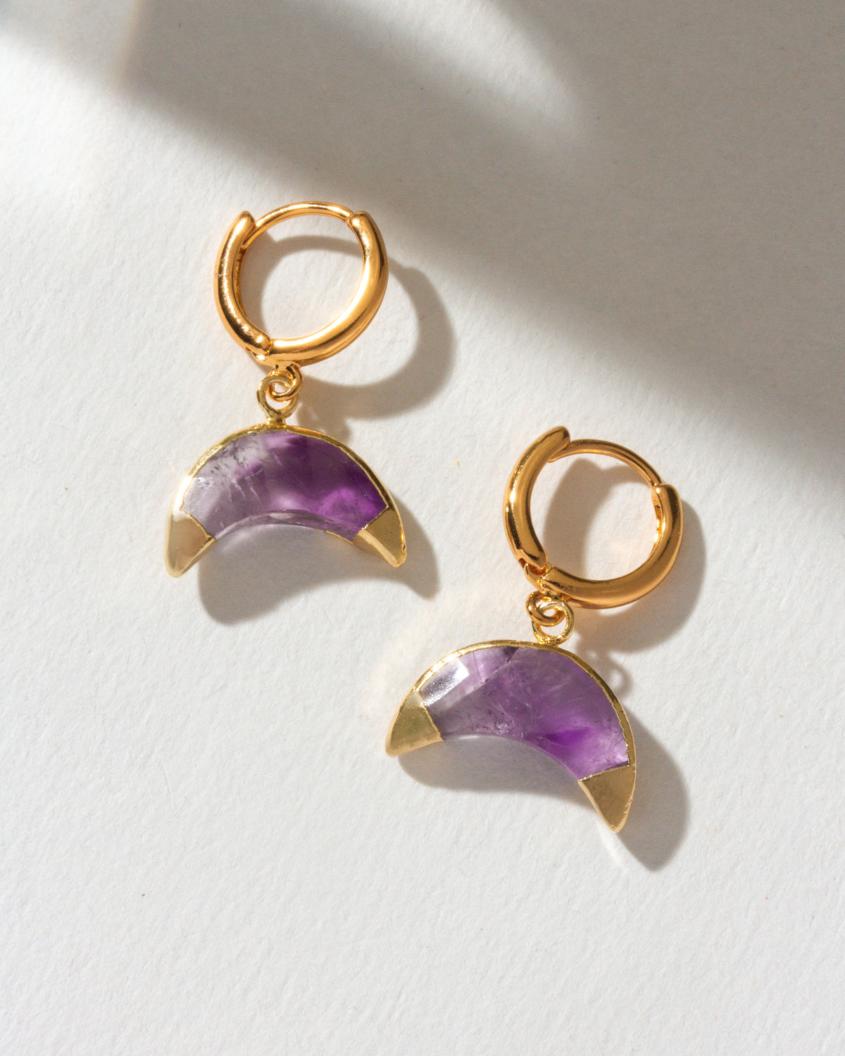 Two gold Crescent Horn Huggie Hoop Earrings with Amethyst gemstones on a white background.