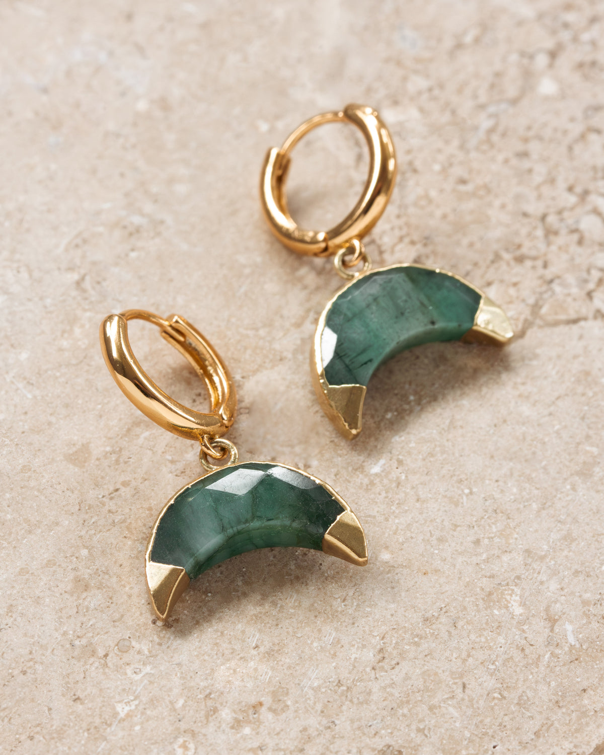 Gold hoop earrings with green crescent moon charms on a beige surface