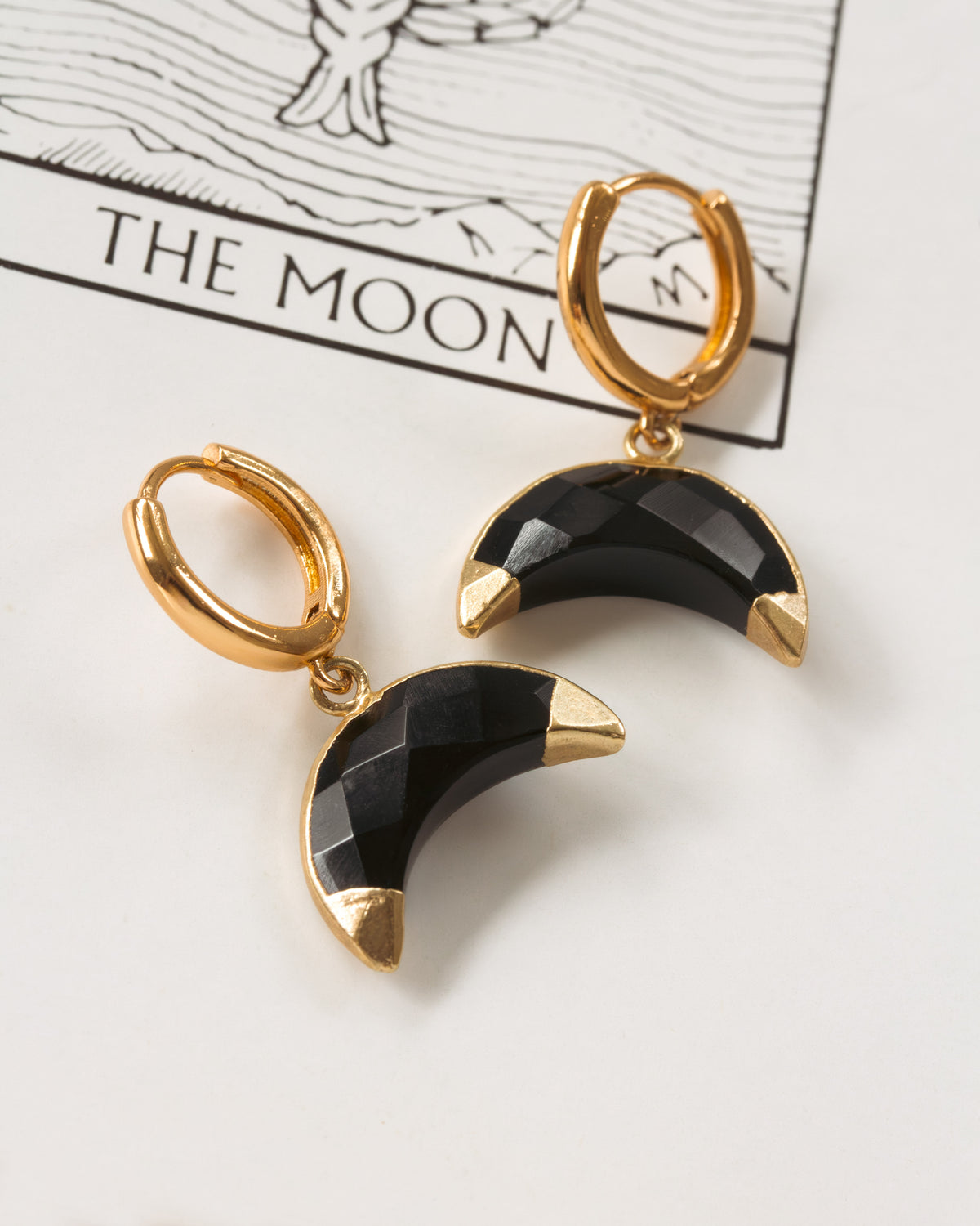 Gold hoop earrings with black crescent moon designs on a tarot card background.