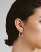 Close-up of a person wearing a gold earring with a red ruby gemstone on a neutral background