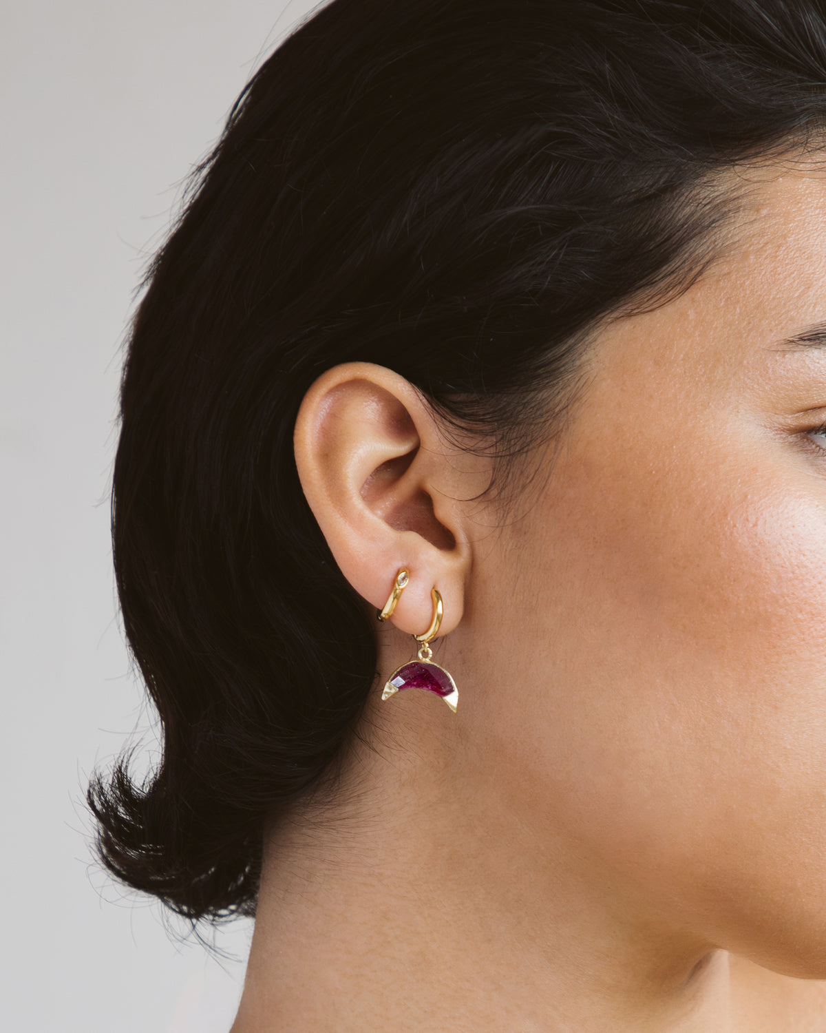 Close-up of a person wearing a gold earring with a red ruby gemstone on a neutral background
