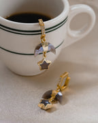 A pair of gold-colored hoop earrings with hexagon and crescent horn charms, encrusted with clear stones and featuring a coffee moonstone star, displayed next to a cup of coffee.