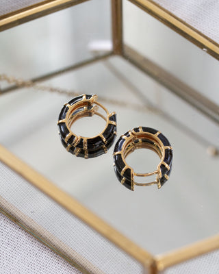 Black striped enamel and gold huggie hoop earrings with gold accents on a glass surface