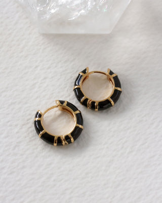 Pair of striped black enamel and gold huggie hoop earrings on a white textured surface