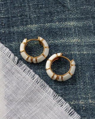 Pair of gold and white striped enamel huggie hoop earrings on a textured blue fabric background