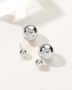 Two pairs of shiny, silver, orb-shaped earrings are displayed on a white surface.