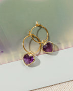 A pair of gold hoop earrings with heart-shaped purple amethyst pendants.