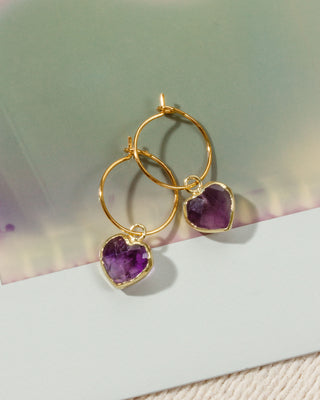 A pair of gold hoop earrings with heart-shaped purple amethyst pendants.