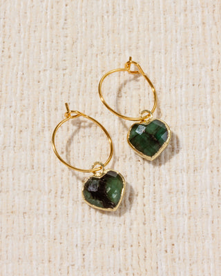 A pair of gold hoop earrings with emerald gemstone heart charms.
