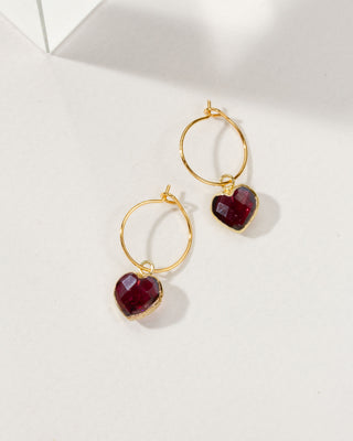A pair of gold hoop earrings with faceted dark red garnet heart-shaped dangling charms.