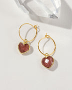 A pair of gold hoop earrings with faceted heart-shaped pinkish-red strawberry quartz charms.