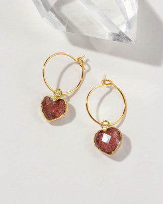 A pair of gold hoop earrings with faceted heart-shaped pinkish-red strawberry quartz charms.
