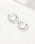 A pair of silver huggie hoop earrings are displayed on a white surface.