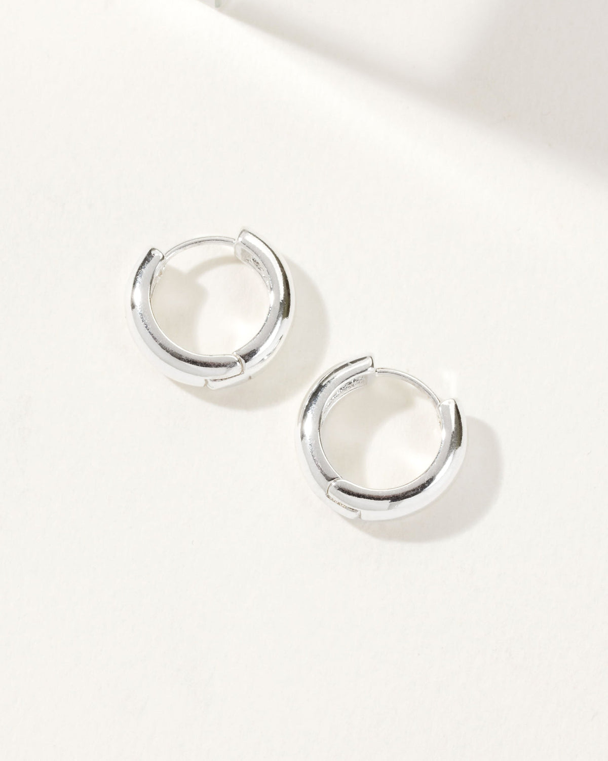 A pair of silver huggie hoop earrings are displayed on a white surface.