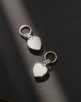 Two silver heart-shaped earring charms with a white textured druzy stone on a dark surface.