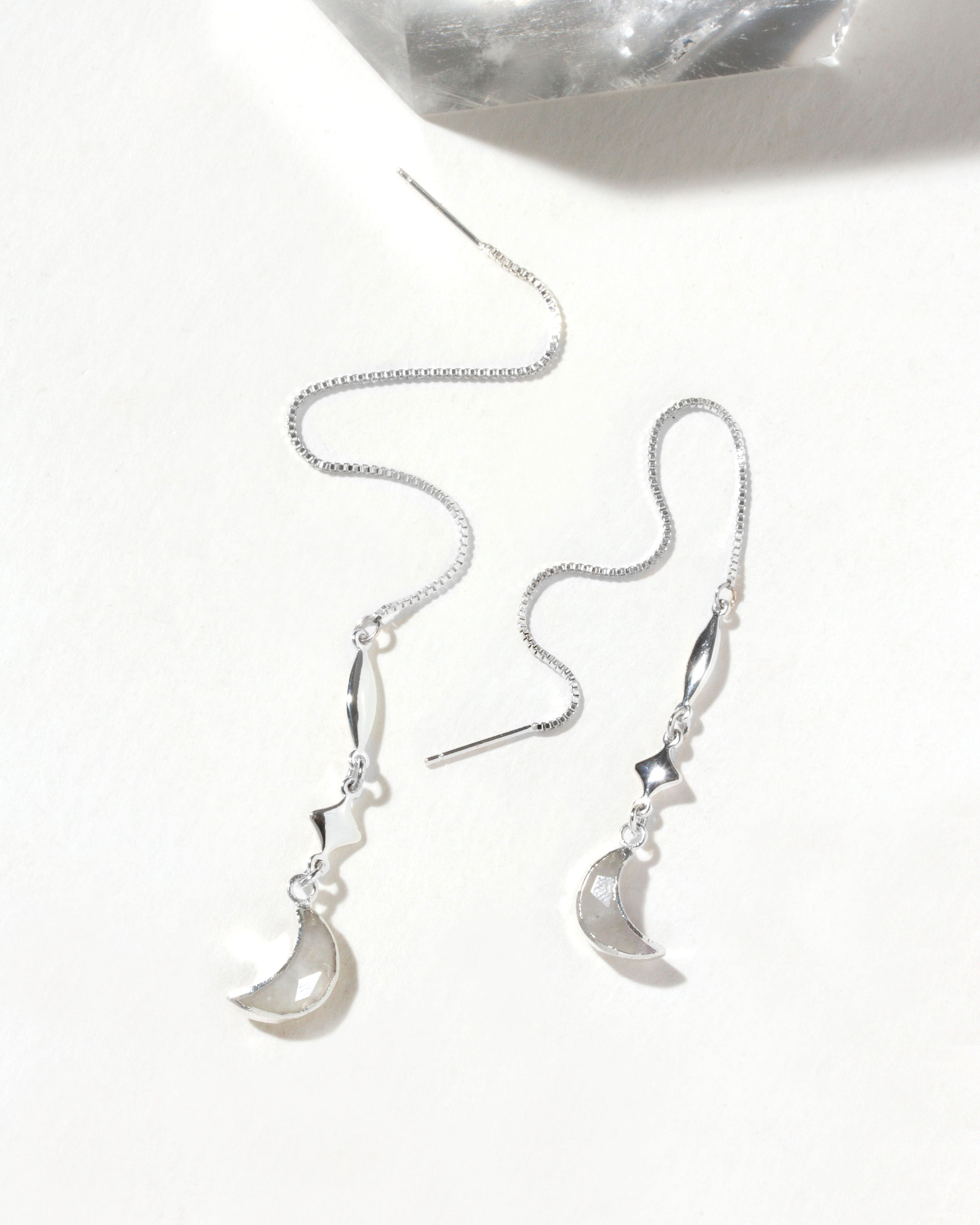 Silver threader earrings with star and moon charms are displayed on a white surface.