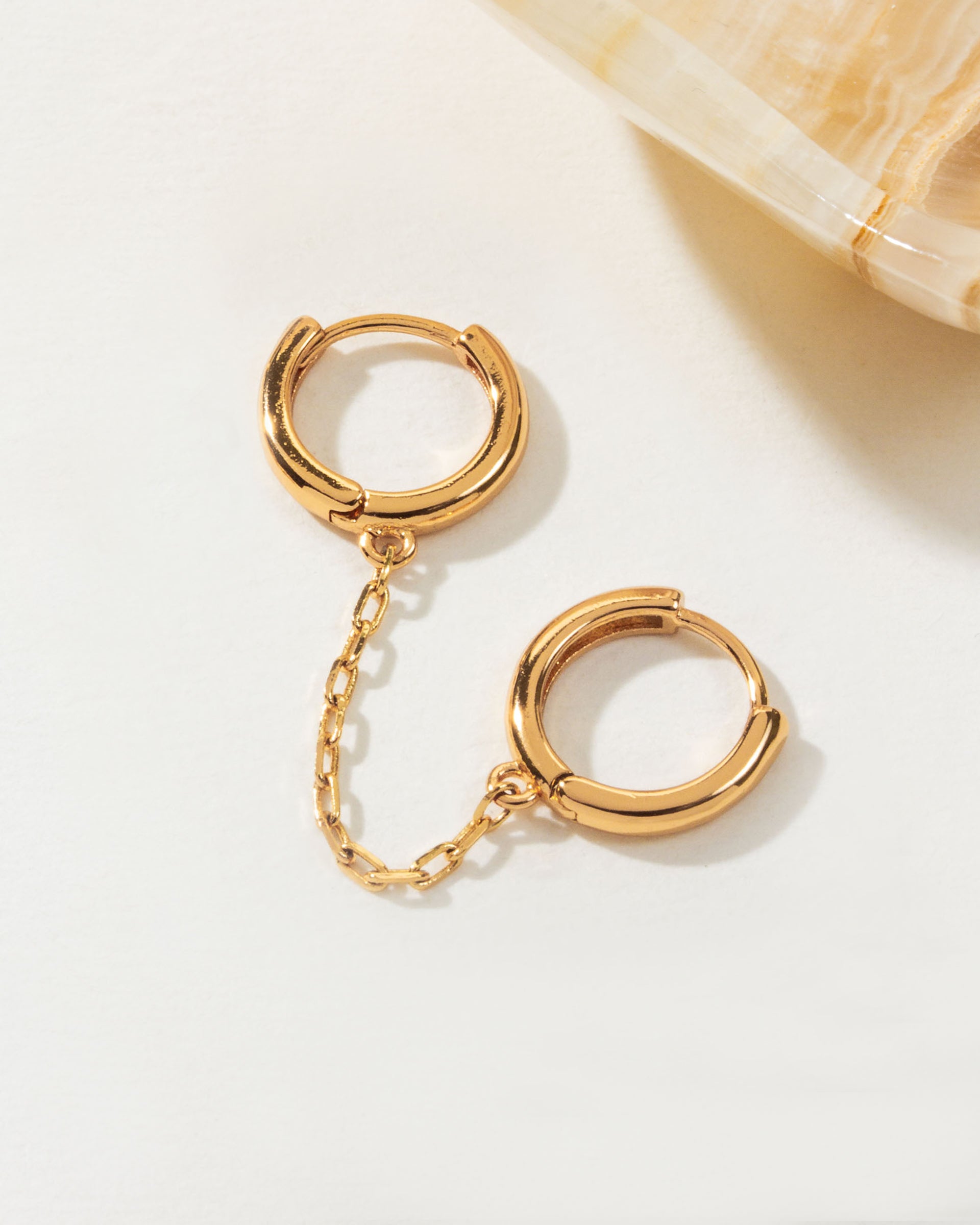 Gold cuffed double pierced huggie hoop earring linked by a chain on a white surface.