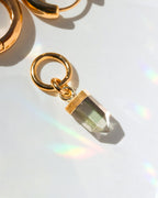 Gold earring single charm with a clear rainbow gemstone on a reflective surface