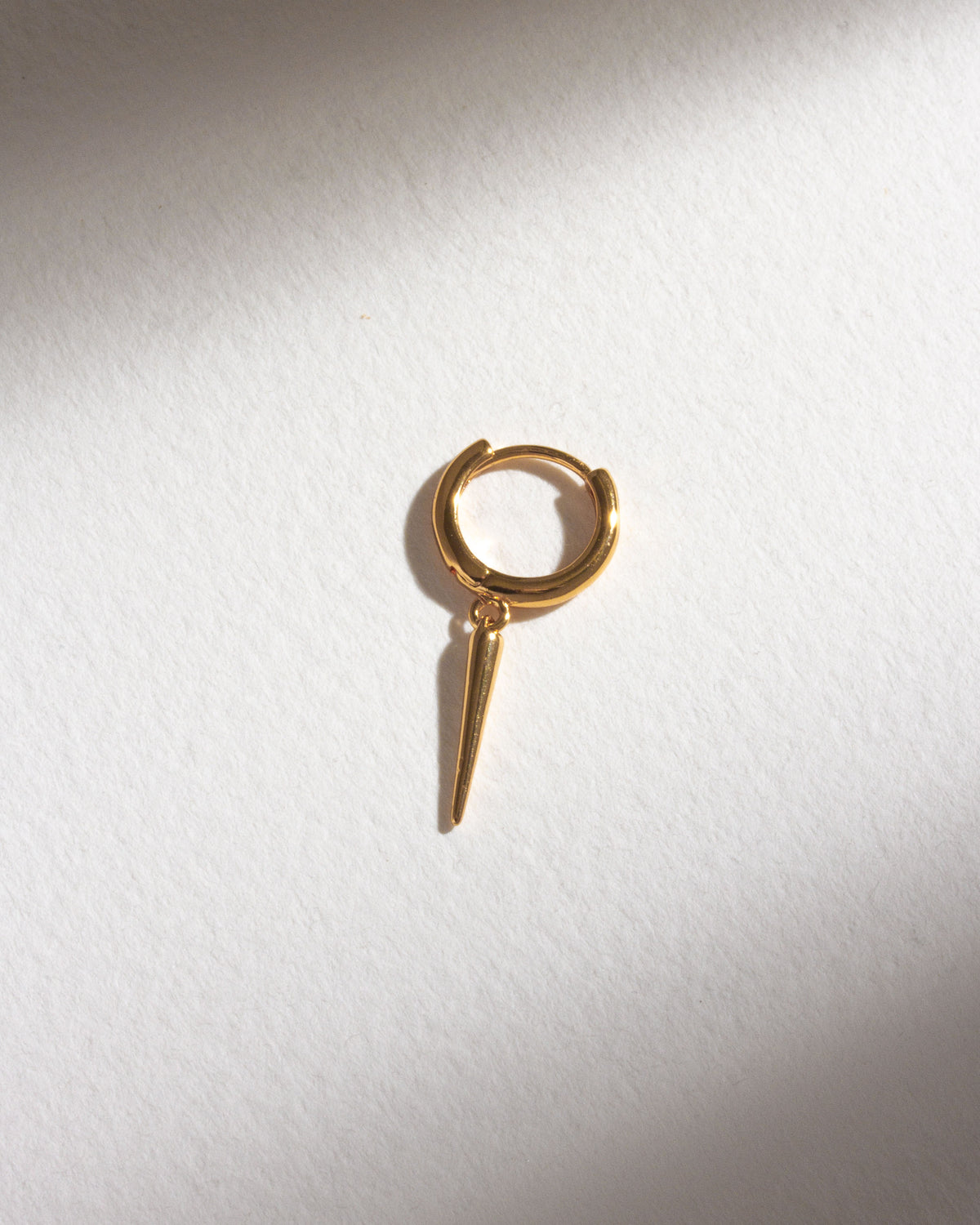 A gold Quill Solo Hoop Earring dangles against a white surface with a shadowed edge.