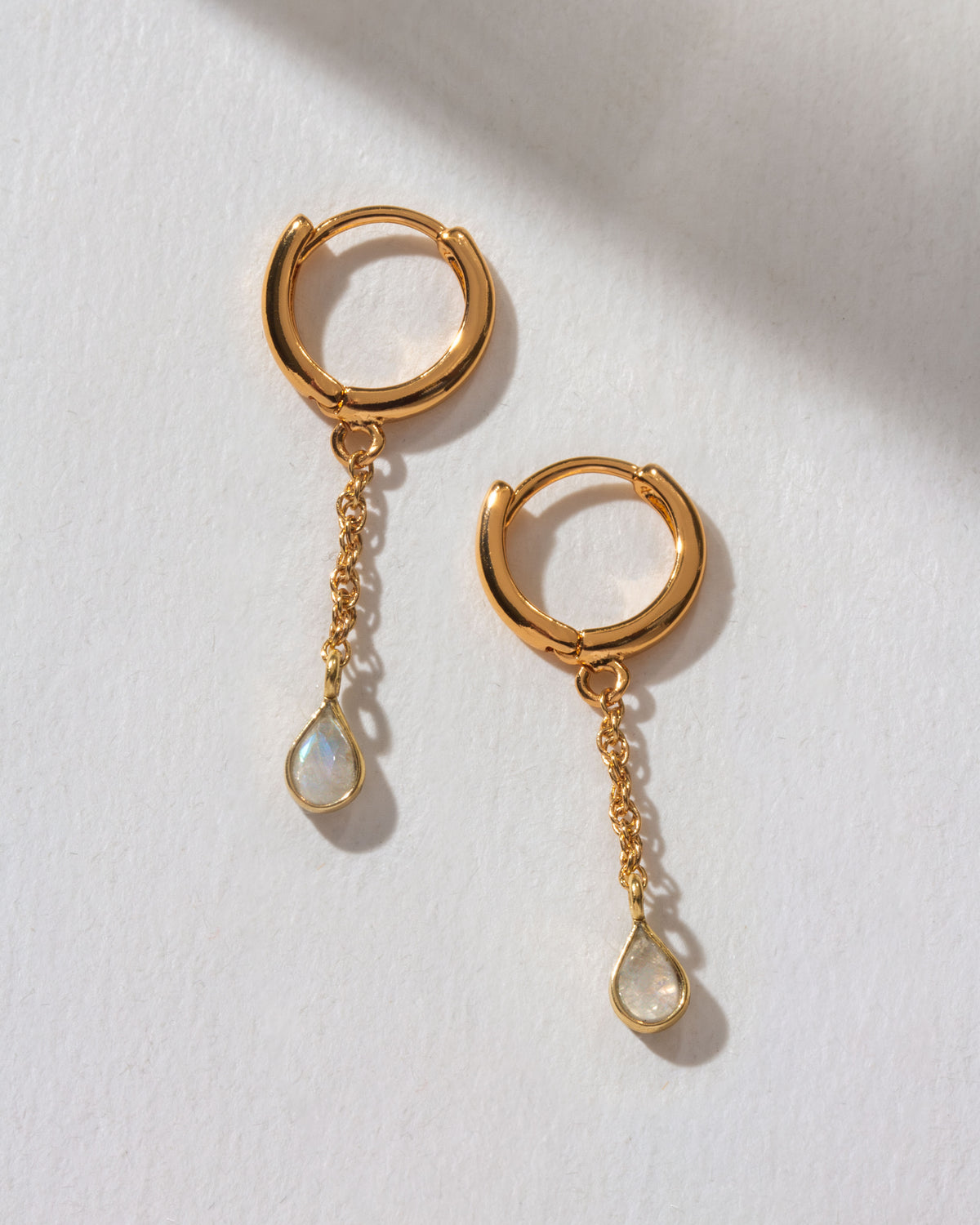 Two gold Misty Huggie Hoop Earrings with Moonstone charms are displayed on a white surface.