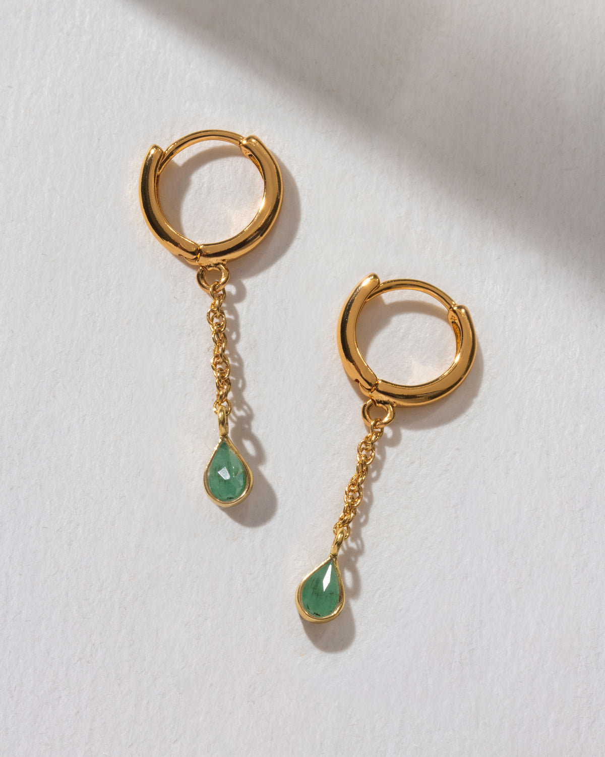 Gold hoop earrings with green emerald teardrop charms dangling from a chain on a white background