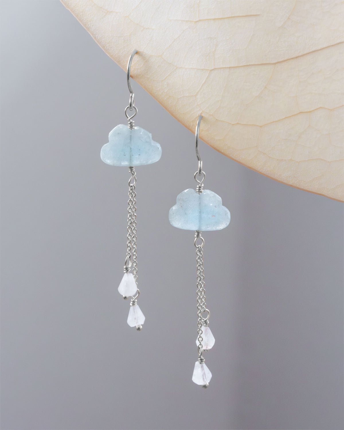 A pair of silver dangle earrings with light blue cloud-shaped gemstone beads and moonstone teardrop beads, hanging against a neutral gray background with a dried leaf overlay.