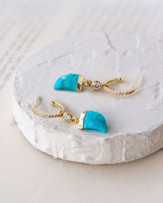 Shark-tooth style gold and turquoise earrings with white crystals on a textured white surface
