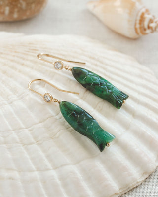 Green Emerald fish-shaped earrings with CZ elements on a textured white surface with seashells in the background
