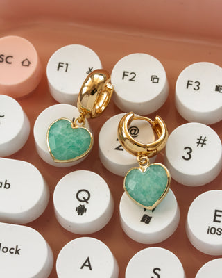 A pair of gold huggie hoop earrings with heart-shaped green amazonite gemstone charms, displayed on a peach-colored keyboard with white keys.