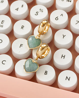 Pair of gold drop earrings with heart-shaped amazonite pendants, on a pink keyboard with white keys.