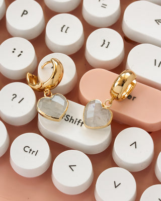 A pair of gold huggie hoop earrings with heart-shaped moonstone faceted gemstone charms, displayed on a pink and white retro typewriter keyboard.