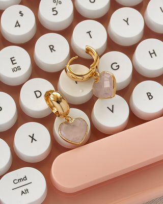 Gold huggie hoop earrings with heart-shaped pink rose quartz stone charms, displayed on a peach-colored keyboard.