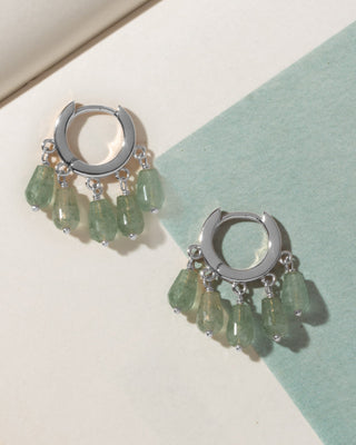 A pair of silver huggie hoop earrings with dangling faceted light green gemstone charms.