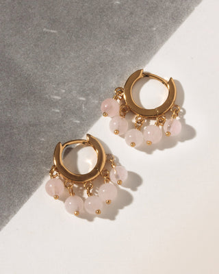 A pair of gold huggie hoop earrings with dangling pink rose quartz beads, displayed on a diagonally split gray and white surface.