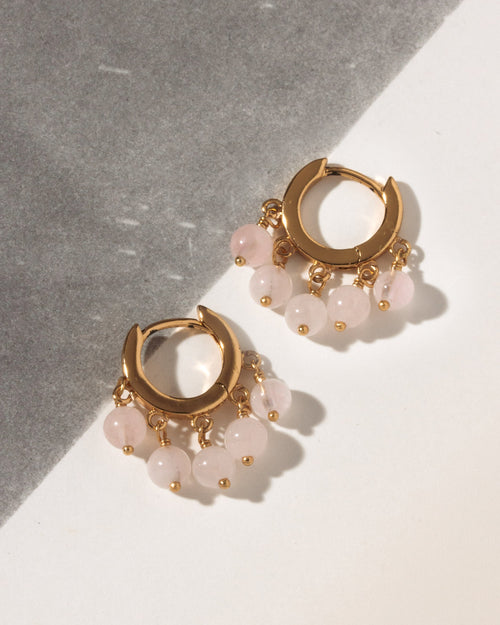 A pair of gold huggie hoop earrings with dangling pink rose quartz beads, displayed on a diagonally split gray and white surface.