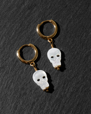 A pair of gold hoop earrings with small white skull charms, displayed on a dark textured slate background.