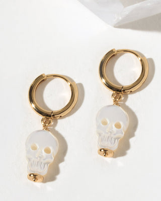 A pair of gold hoop earrings with dangling white mother of pearl skull charms.