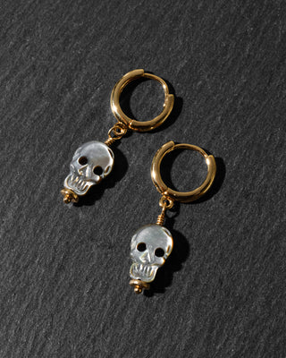 A pair of gold hoop earrings with dangling silver skull charms, displayed on a dark textured slate background.